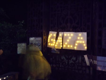 Young woman at vigil looks at homemade sign by Larry Haiven, Gaza in tea lights